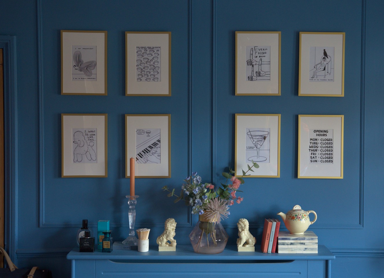 A blue wall, sketched prints, a shelf filled with ornaments and books