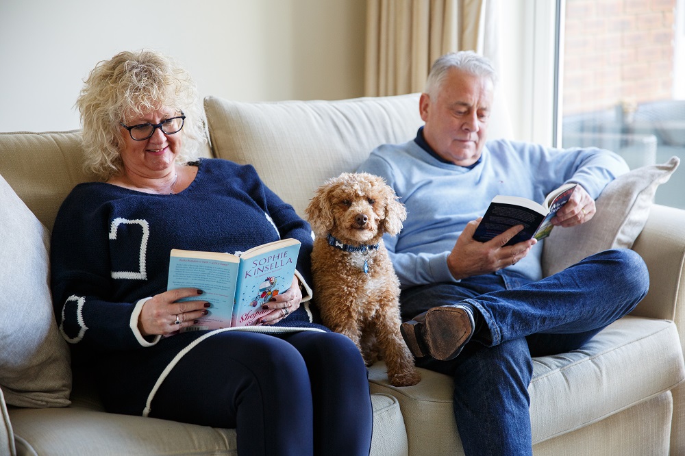 Two people sat on a sofa reading with a dog