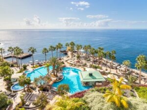 The view from a coastal apartment that looks over two pools, surrounded by palm trees and the azure blue sea